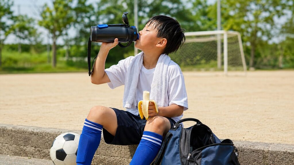 練習後のグラウンド脇で、水筒から水分を取りながらバナナを食べる少年