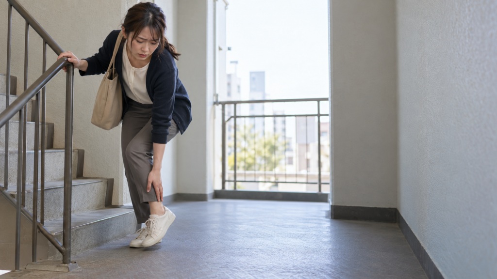 階段の踊り場で手すりにつかまり、痛む足首に手を添えながら立つ女性